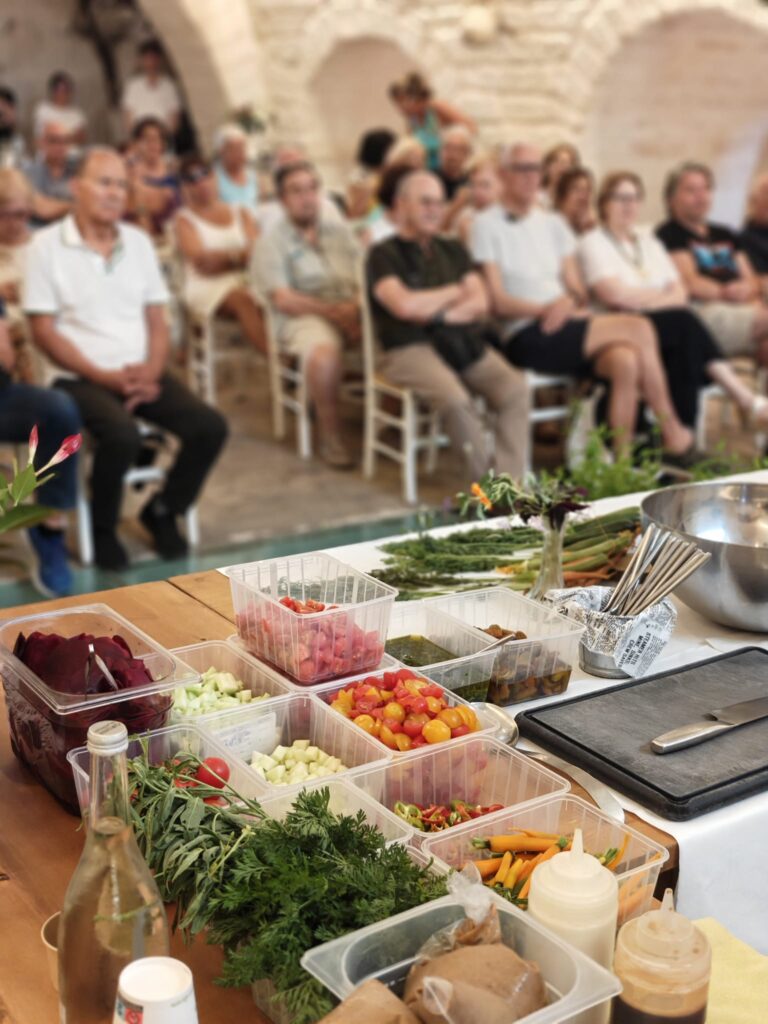 Ingredienti freschi e ortaggi di stagione e biologici disposti su un tavolo da cucina, con il pubblico seduto sullo sfondo durante una lezione a Casale Pontrelli per l'UTE