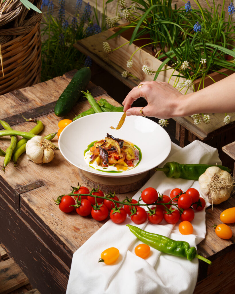 Piatto gourmet con bucce e ortaggi di stagione, servito su crema vegetale, circondato da verdure fresche su tavolo rustico in legno.